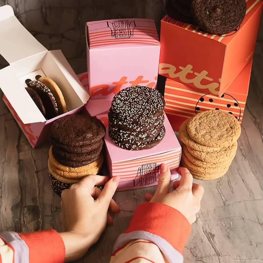 Duas mãos segurando cookies e brownies sobre caixas rosa e laranja.